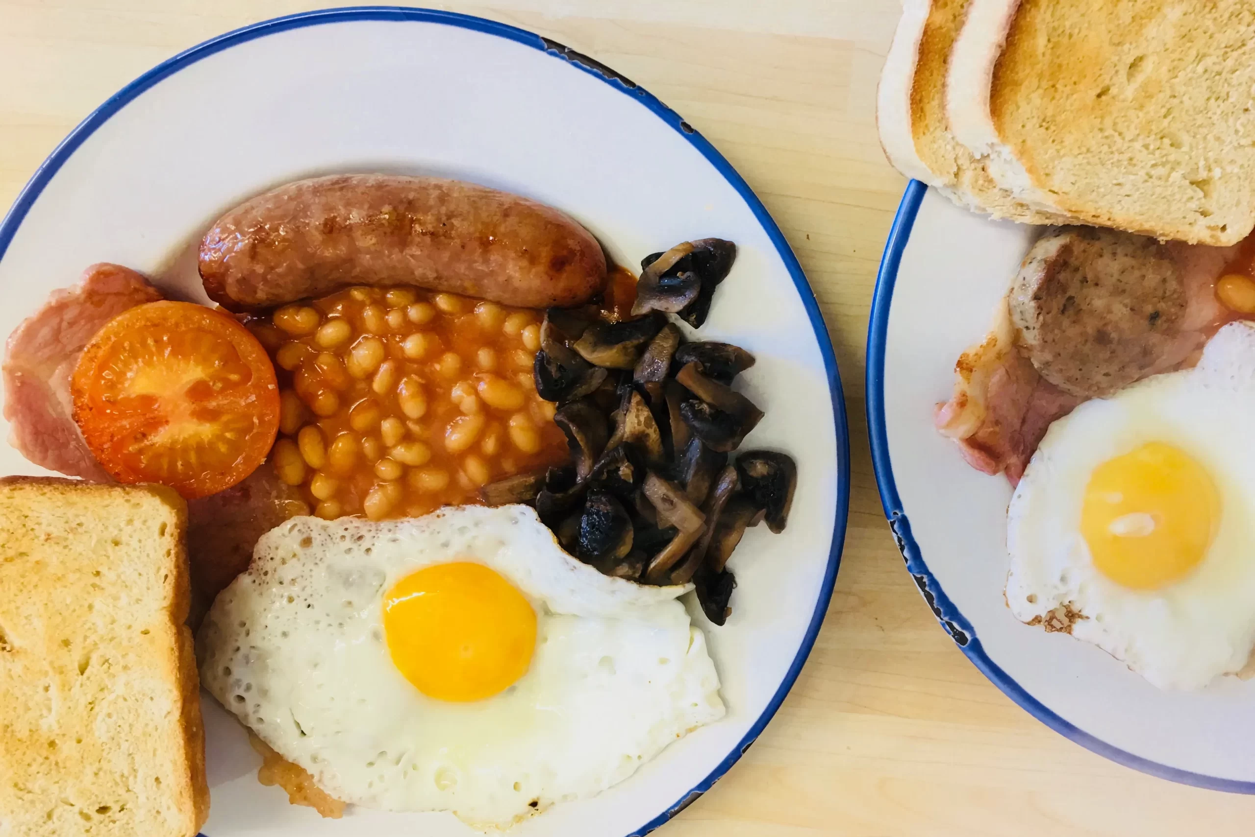 Plates with a full English breakfast: fried eggs, beans, sausage, bacon, tomatoes, mushrooms, and toast grace the light wood table of the cosy breakfast bar. St Georges Perranporth