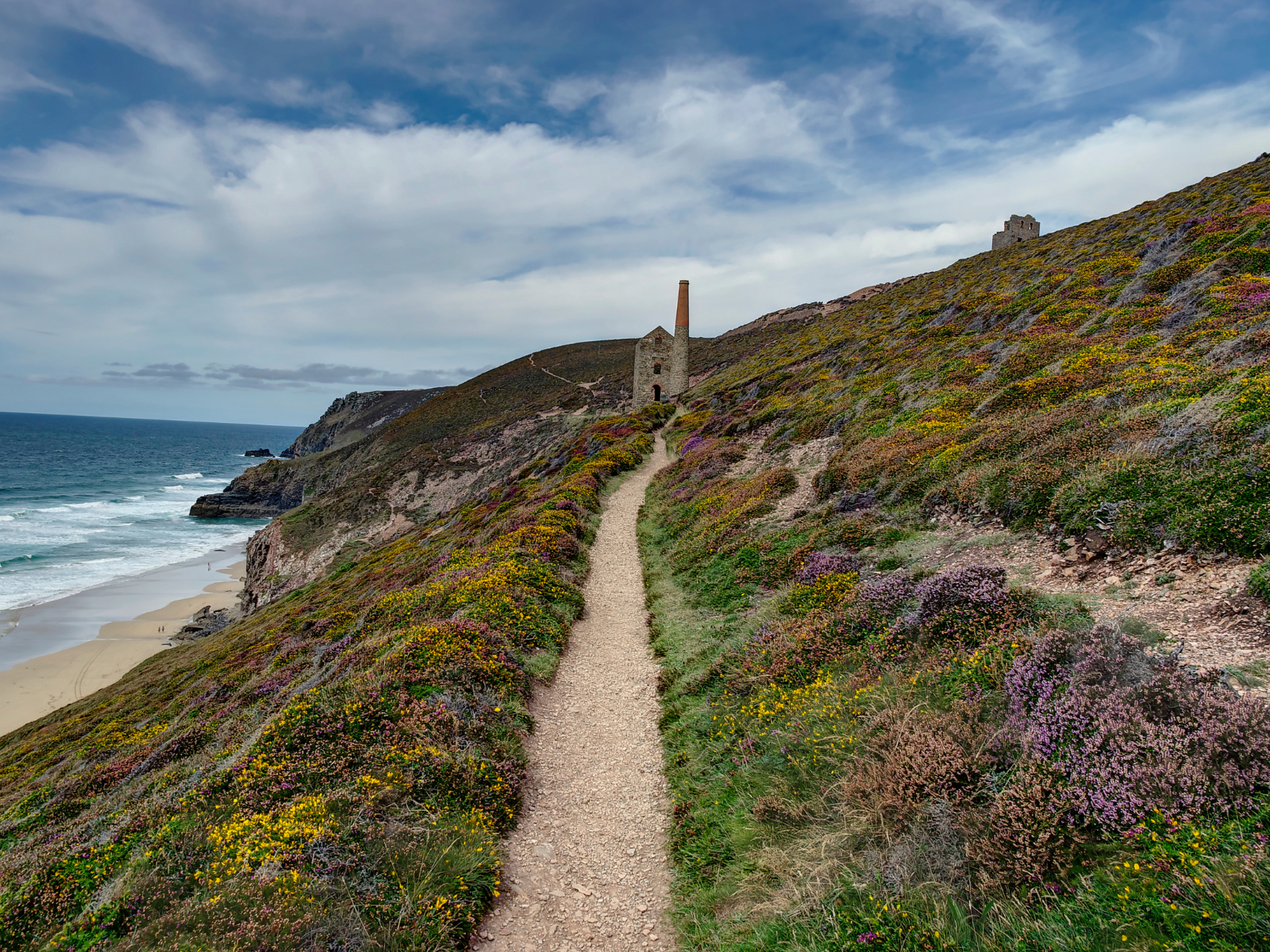 A narrow path leads along a coastal hillside covered in wildflowers, with an old stone building and the ocean visible in the background under a cloudy sky. St Georges Perranporth